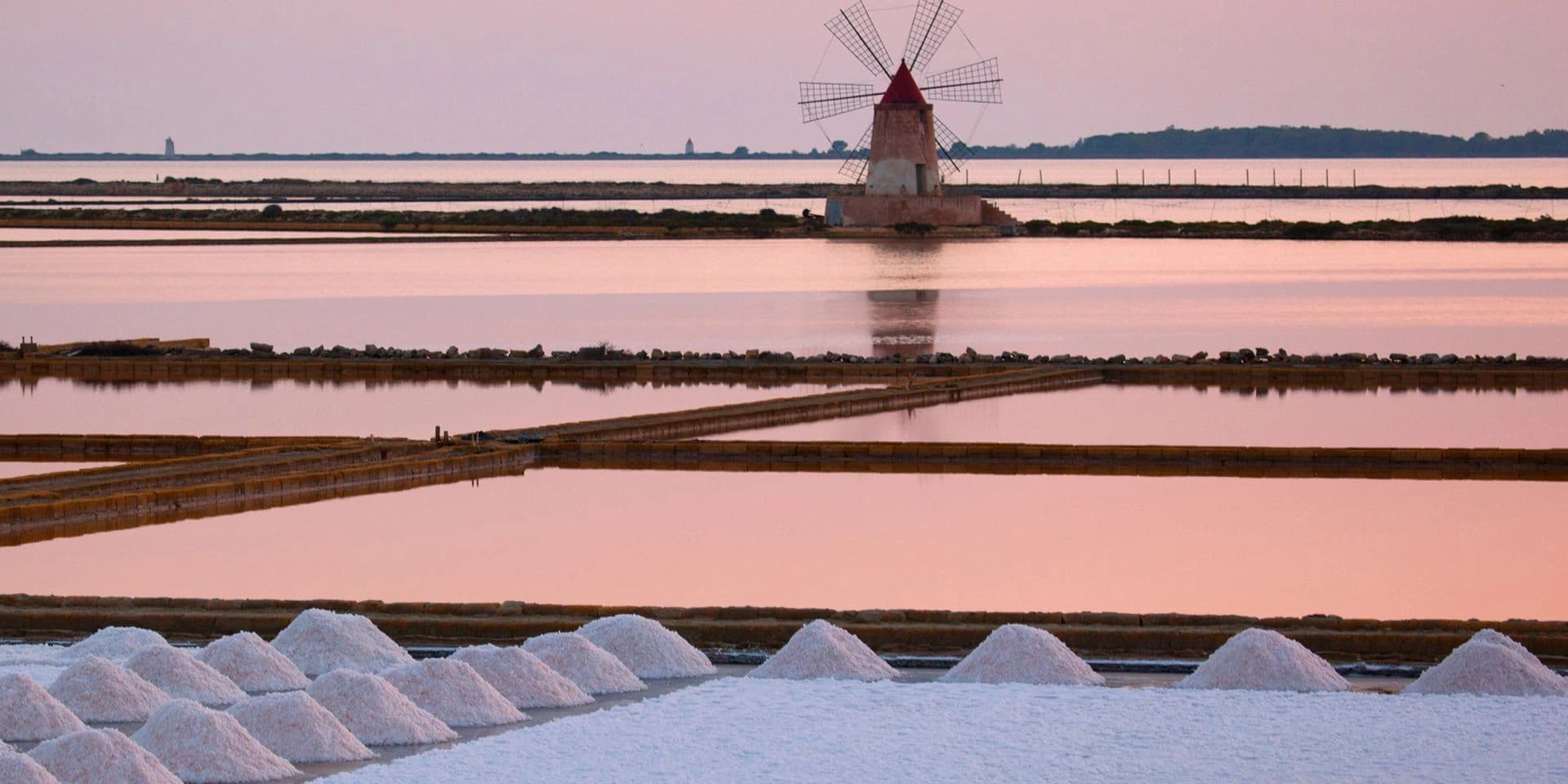 A windmill standing among the salt pans at sunset, its blades dark against an orange sky, with heaps of white salt in the foreground