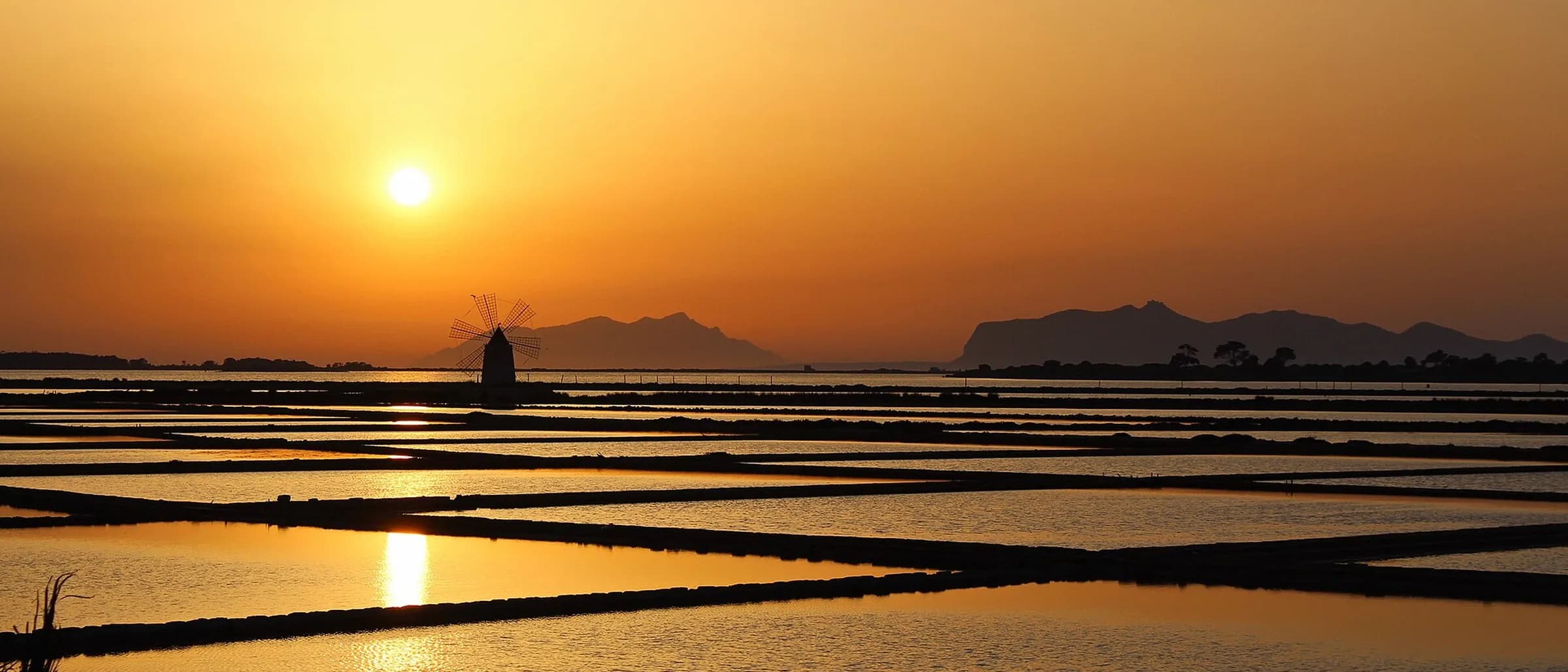 The sun low over the salt pans at sunset, a windmill in silhouette beside the shallow basins with mountains along the horizon