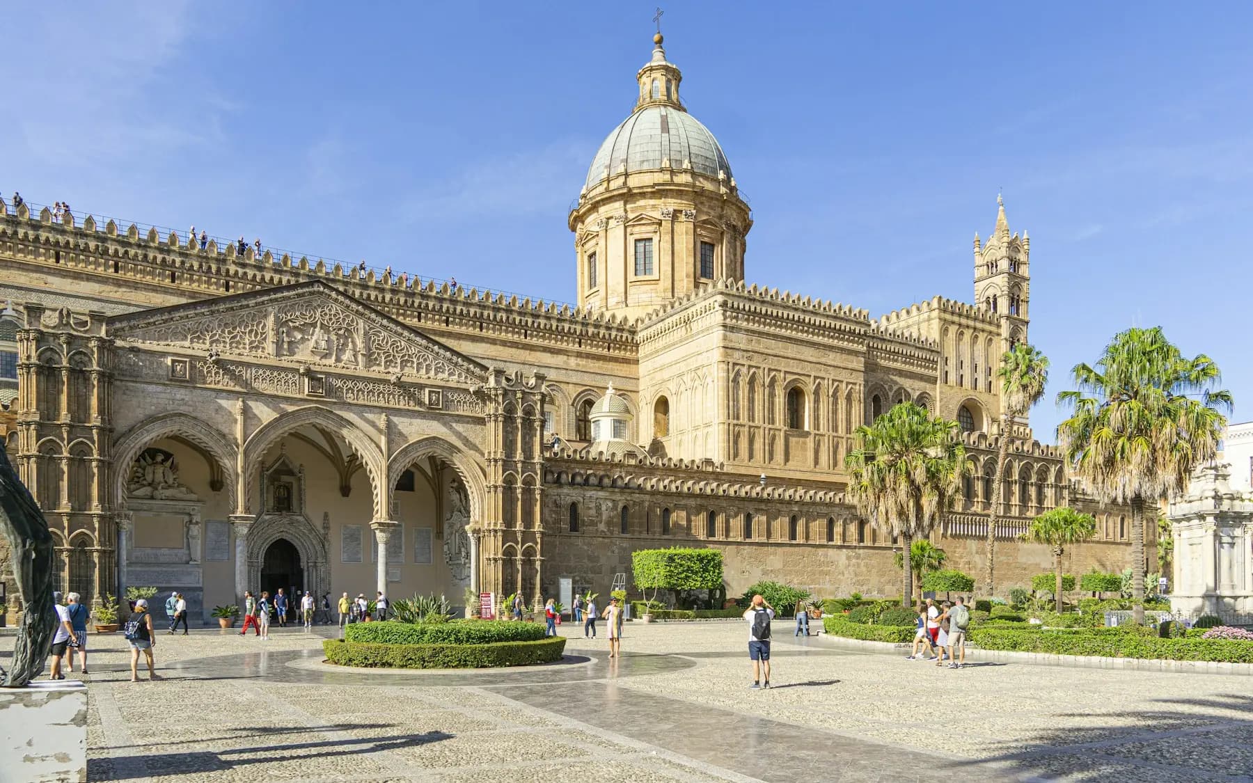 Palermo cathedral seen from its forecourt, with the dome, the arcaded stone porch, palm trees along the side and visitors crossing the square