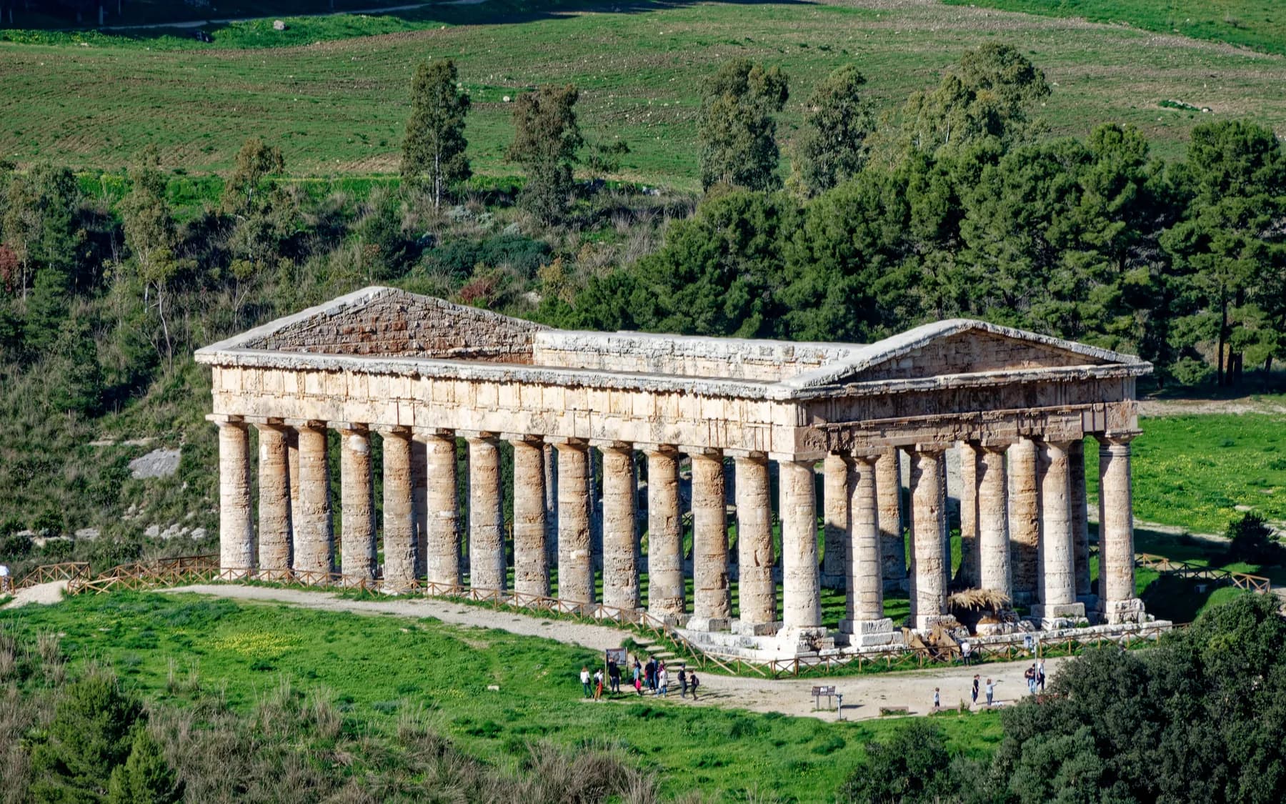 The Doric temple at Segesta seen from above, standing alone among green fields and trees, with visitors gathered at its base